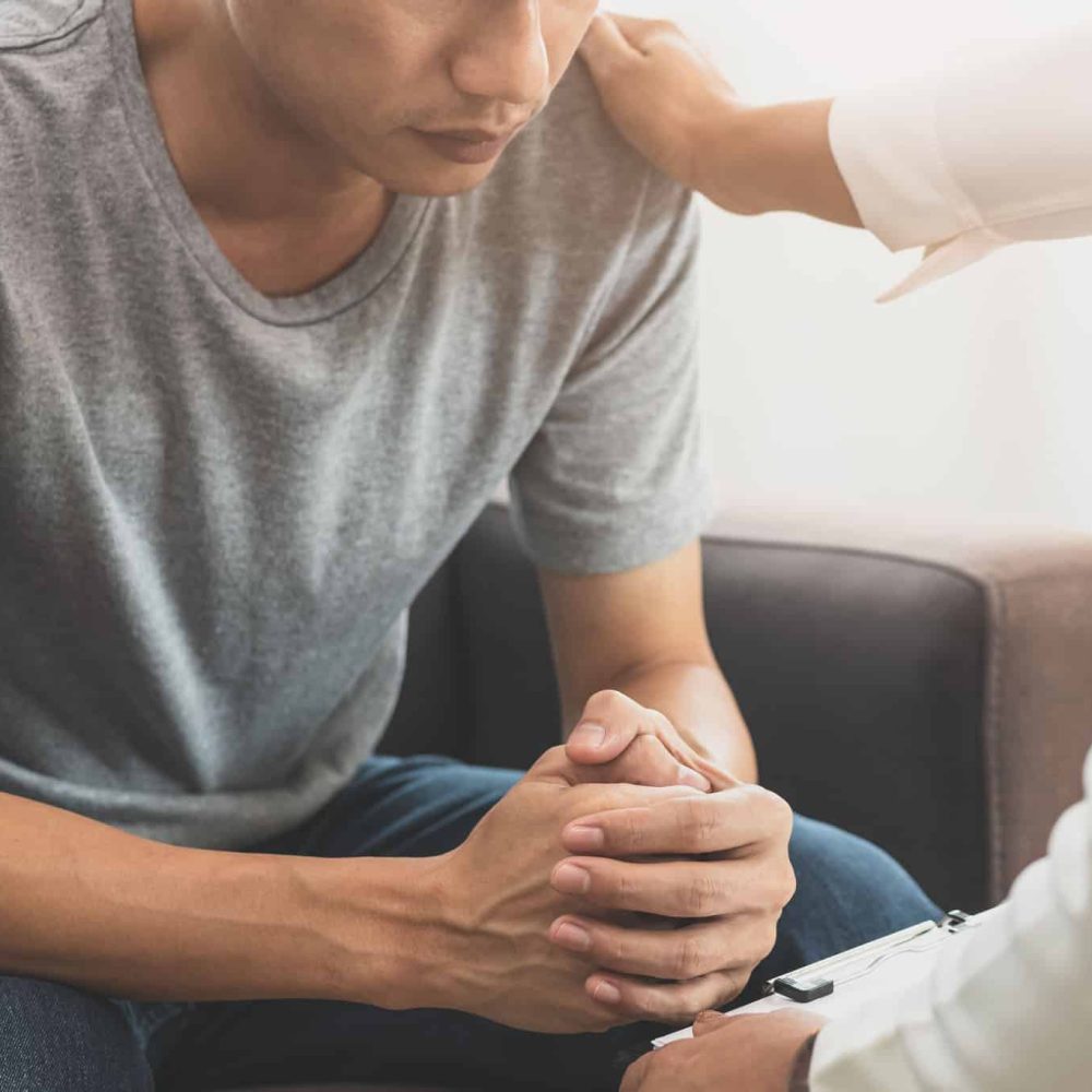 Young asian man, male suffers from a mental who needs to therapy with a psychologist while sitting on couch to consult, psychiatrist has encouragement the patient by touching to make his feel relaxed.