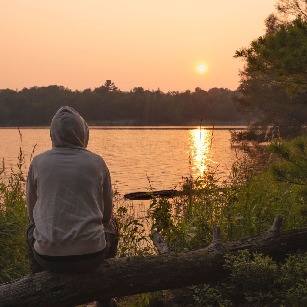 Man wearing hoodie sitting alone at lookout on the beach by calm lake watching sunset and orange sky at rural area. Camping, hiking, portaging, active lifestyle concept.